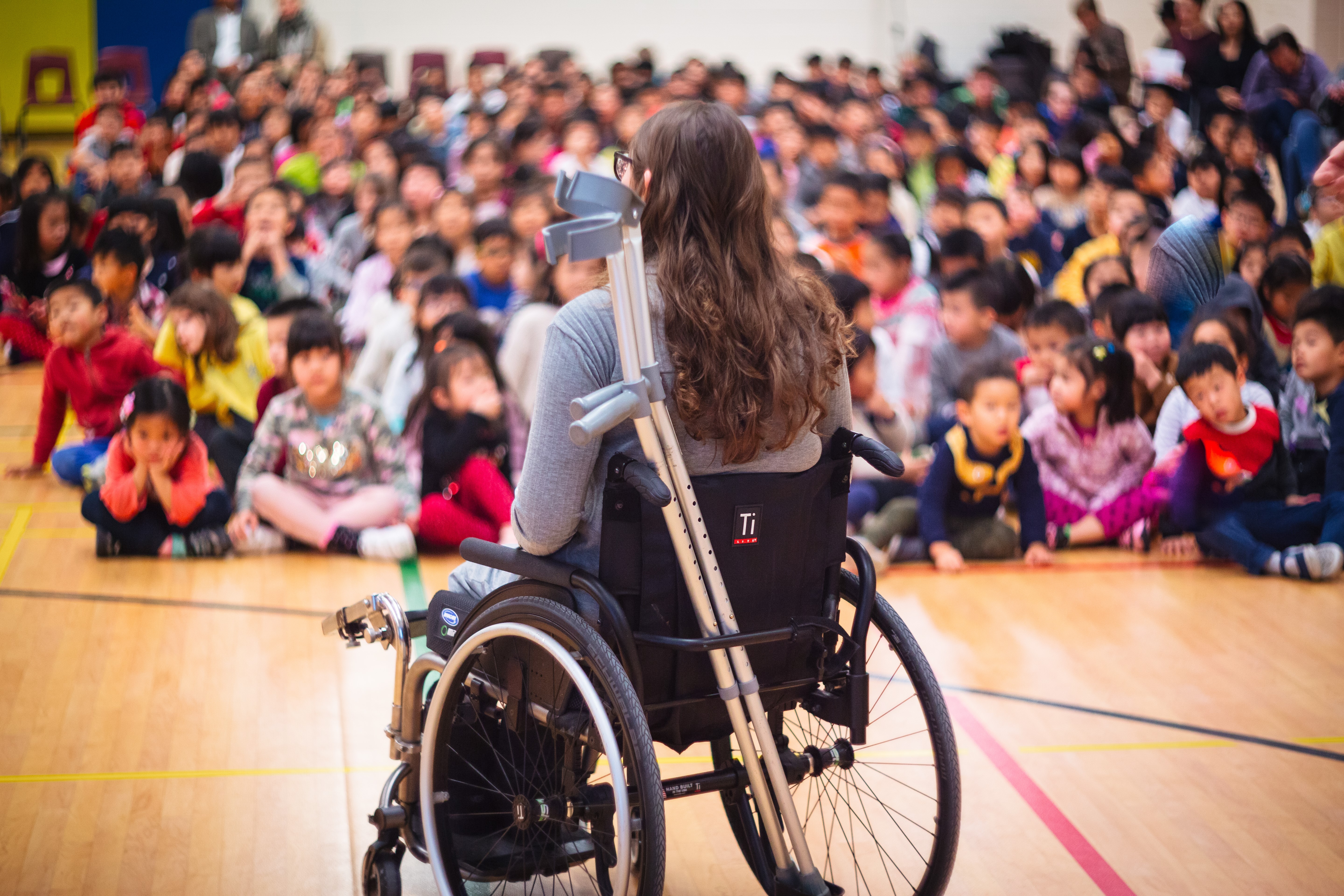 woman in a wheelchair from behind, speaking to a group of children in a gymnasium, out of focus